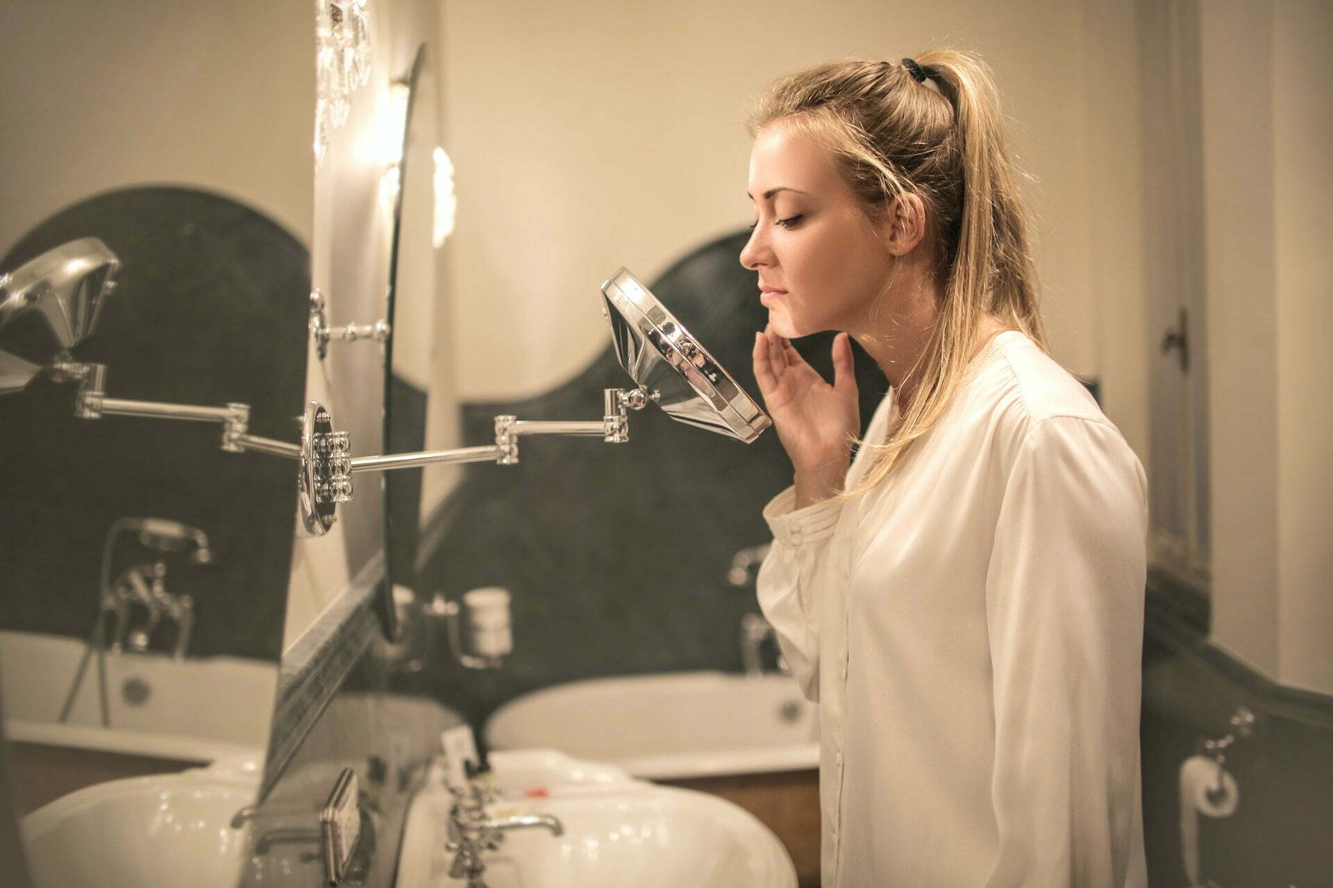 Woman touching her chin and looking at her face in a round mirror in a bathroom, considering how to manage acne.