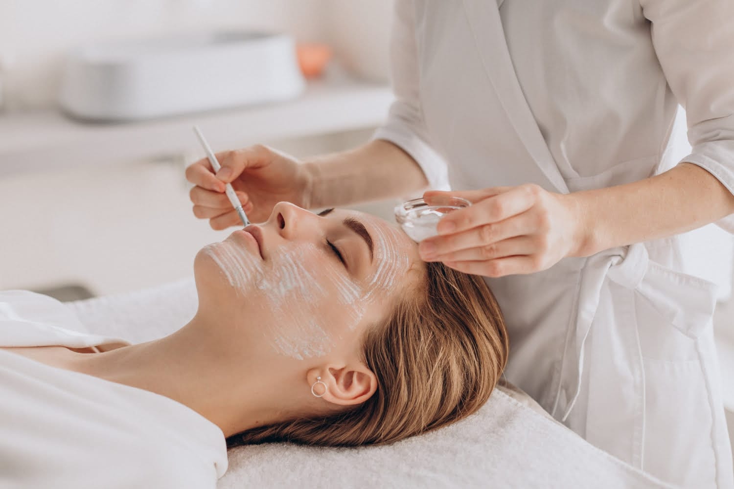 A person applies a facial mask to a woman lying on a spa treatment table, showcasing the relaxing benefits of professional facial treatments.