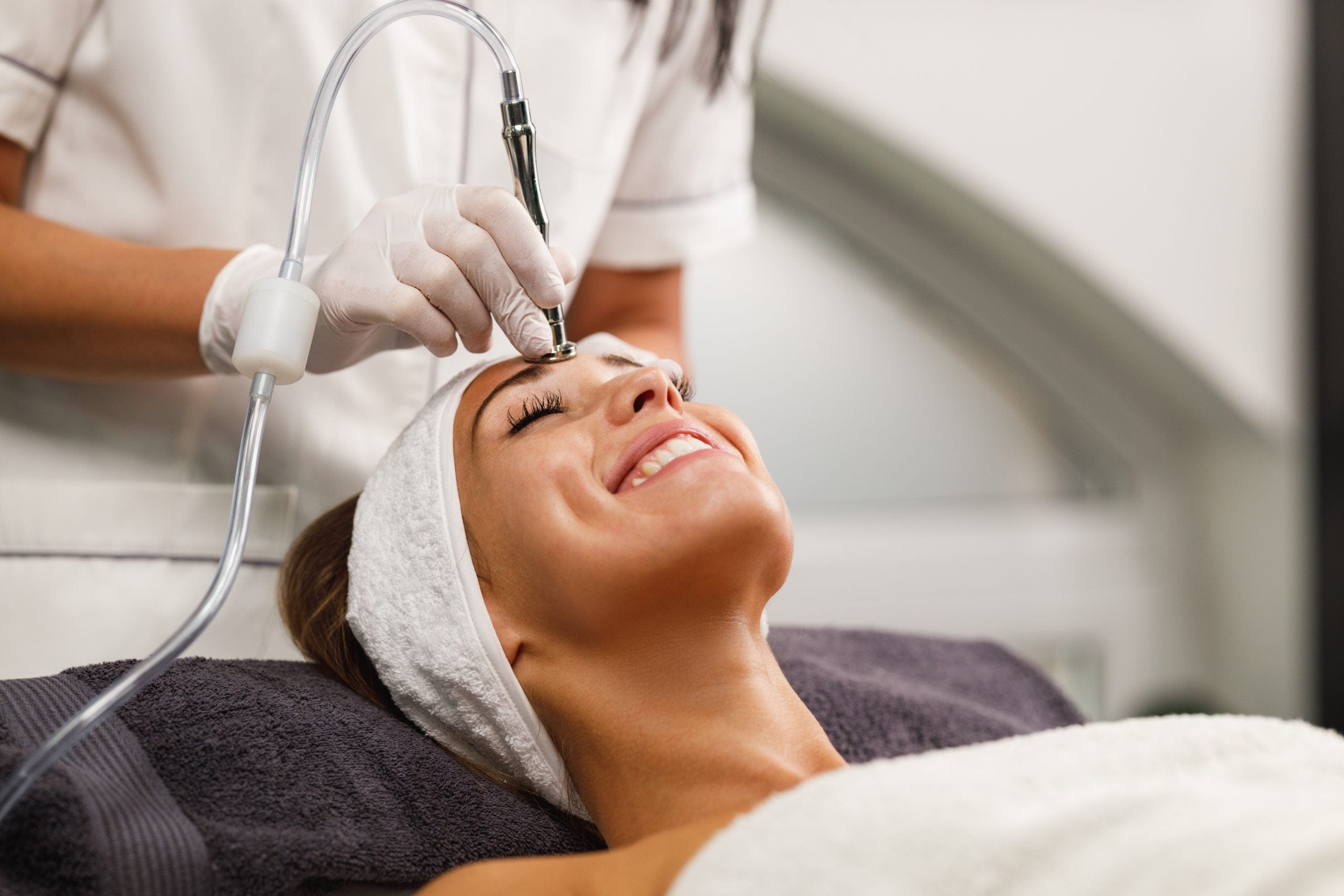 A woman smiles while receiving a hydrodermabrasion facial treatment from a professional using a skincare device.