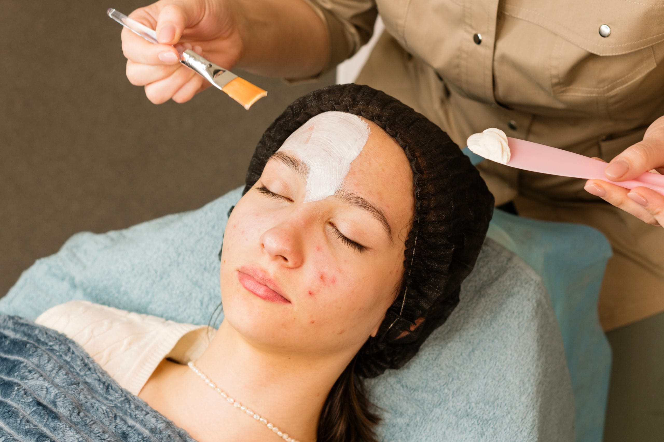 Person with closed eyes receiving a facial mask applied to their forehead with a brush at a spa.