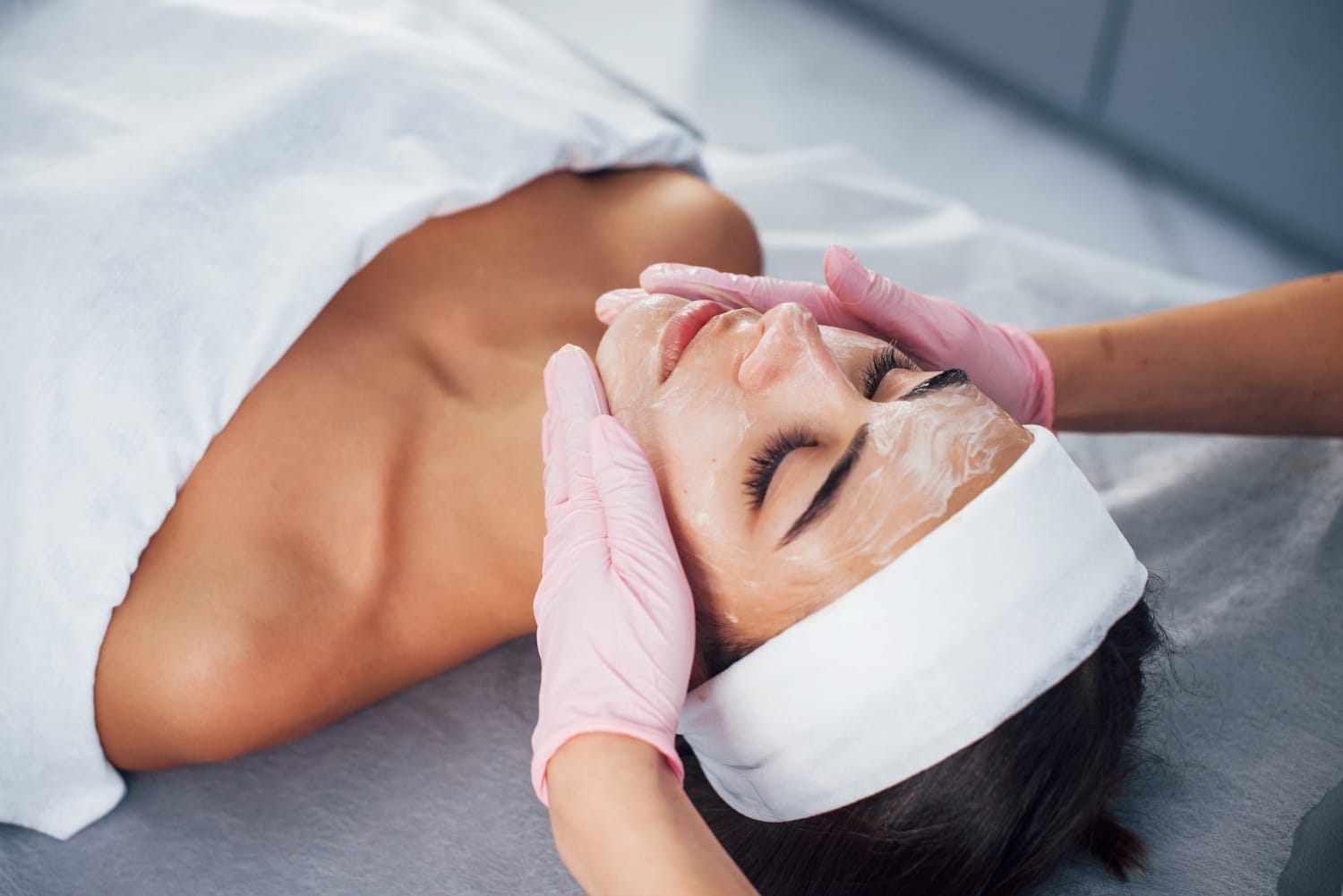 A woman with a headband receives a facial treatment with cream applied by hands in pink gloves.