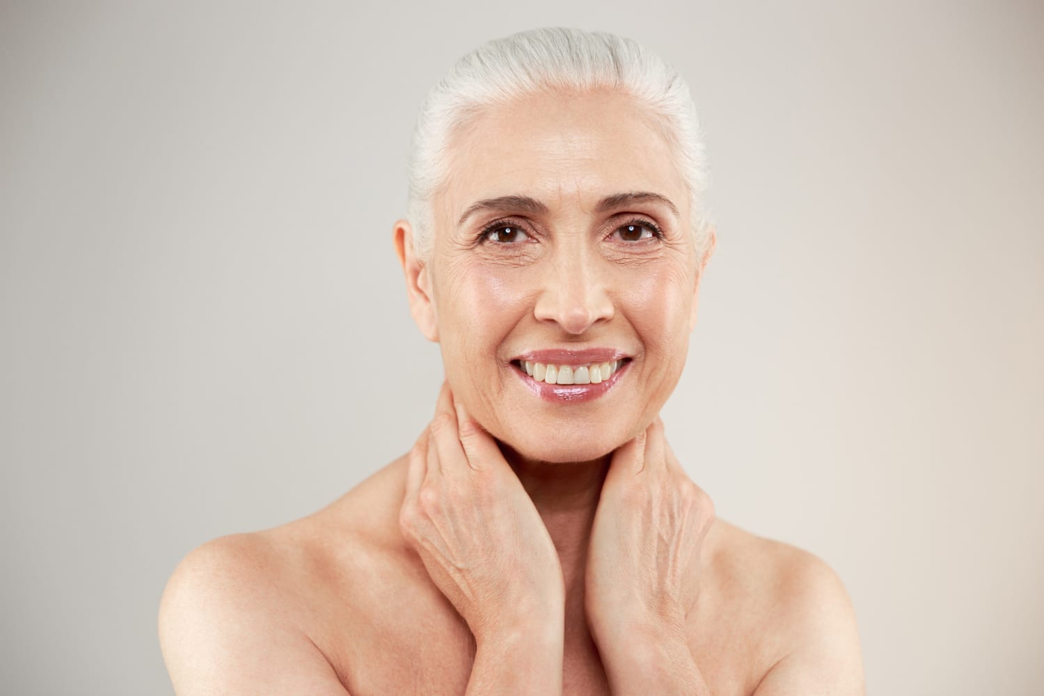 Smiling older woman with gray hair touching her neck, posing against a light gray background.