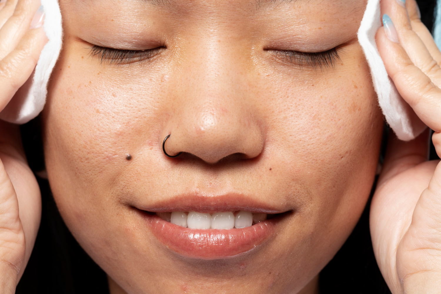 Woman with closed eyes smiling while holding cotton pads to her face, close-up view of clear skin.