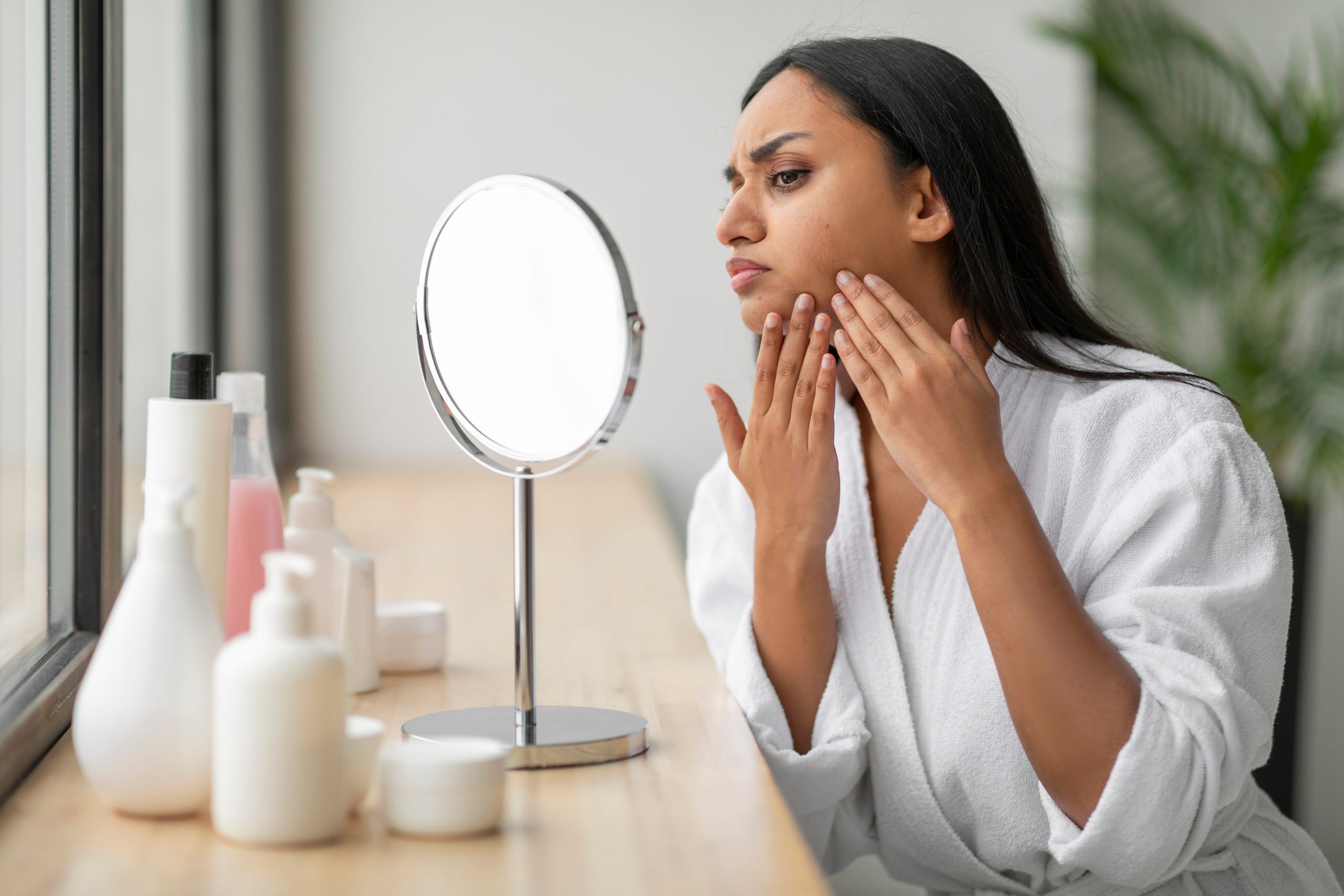 Upset young indian woman looking at mirror, touching her face Woman in a white robe examines her face in a mirror, touching her cheek with a concerned expression.